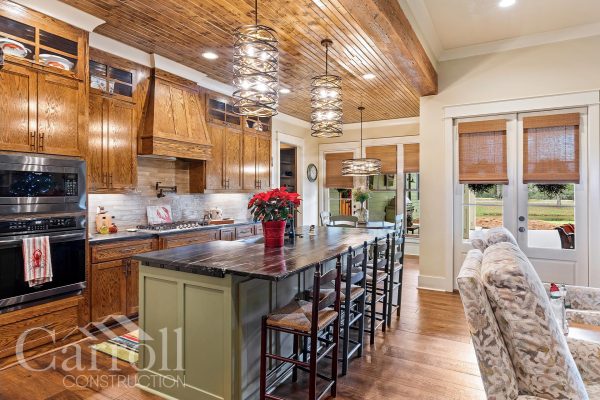 another image of custom kitchen with stained oak cabinets, pine ceilings and modern chandeliers. Undercounter lighting displaying a linear pattern backsplash near a black soapstone.
